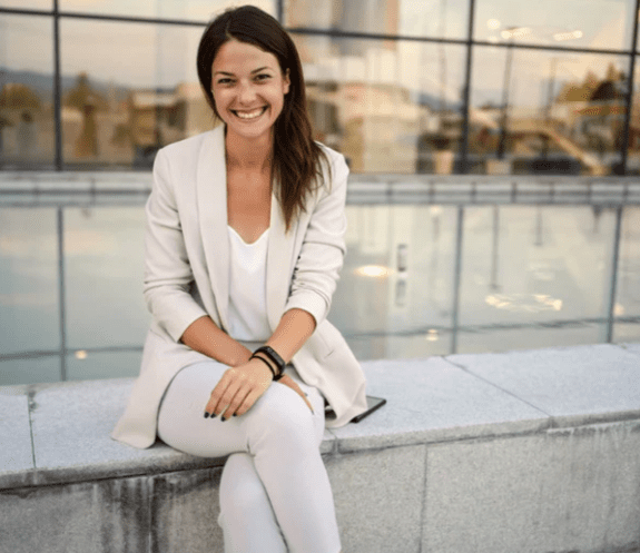 Female sat on concrete wall. There's an office building behind her. She is wearing all cream with trousers top and jacket. She has dark hair mid length and wavy. She is smiling at the camera. Her hands are crossed on her lap.